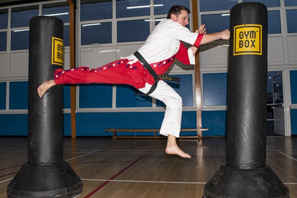 A Taekwondo practitioner in mid-air executing a flying side kick at a standing punching bag in a gym.